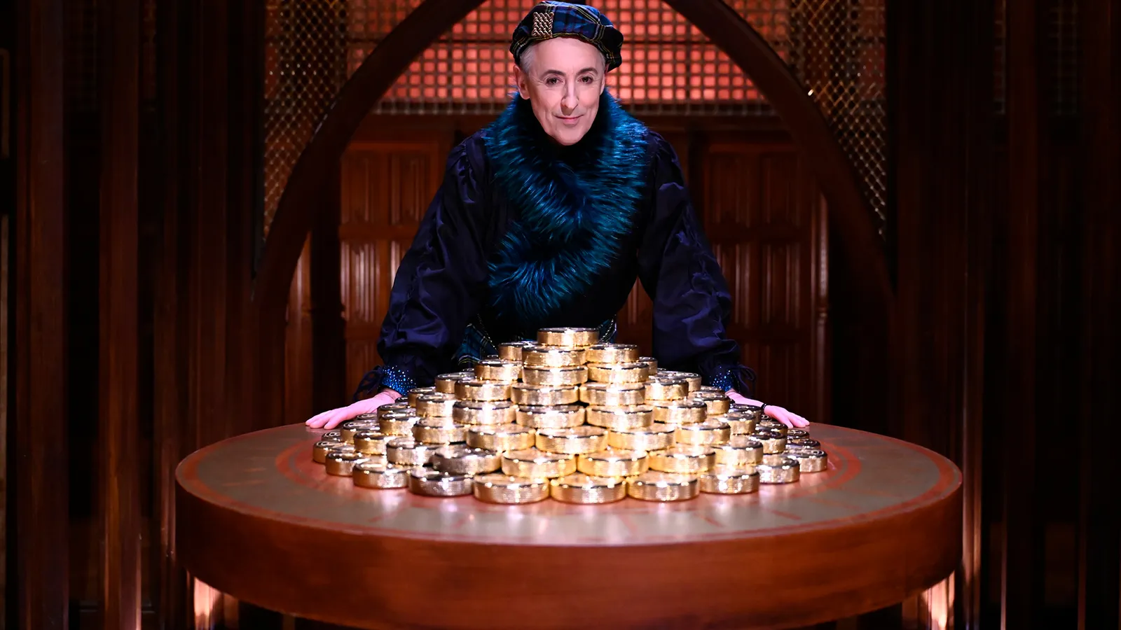 Alan Cumming behind a round table heaped with gold bars