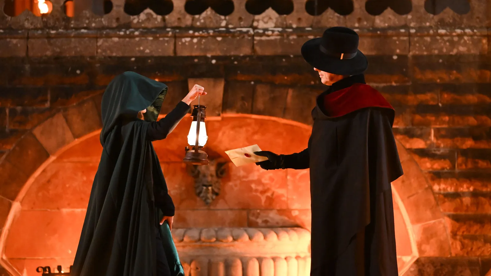 A hooded figure holding a lantern takes a sealed envelope from Alan Cumming at night