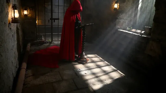 A hooded figure in a red cloak in a shaft of light in the castle