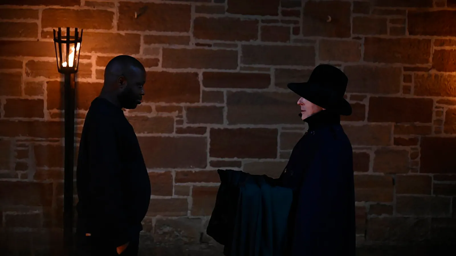 Alan Cumming offers a dark cloak to a player face to face against a stone wall at night
